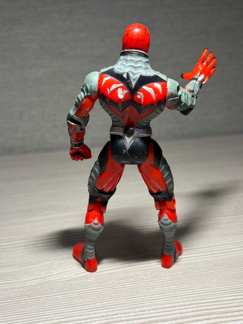 May include: A red and gray action figure with a detailed design. The figure has a red helmet, red hands, and red boots. The back of the figure features a black and red design with gray accents. The figure is posed with one arm raised.
