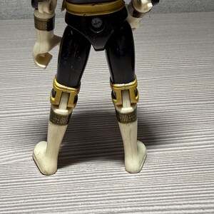 May include: A vintage action figure, seen from the rear, in black and gold. The figure has white limbs and gold detailing on the joints and armour. The text "&copy; 96 BANDAI MADE IN CHINA" is visible on the figure's back.