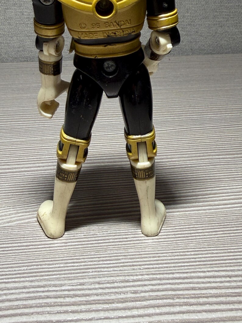 May include: A vintage action figure, seen from the rear, in black and gold. The figure has white limbs and gold detailing on the joints and armour. The text "&copy; 96 BANDAI MADE IN CHINA" is visible on the figure's back.