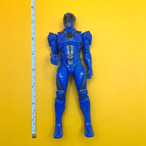 May include: A blue superhero action figure, standing on a yellow surface. The figure is approximately 25 cm tall, with detailed armor and a helmet. A measuring tape is positioned next to the figure.