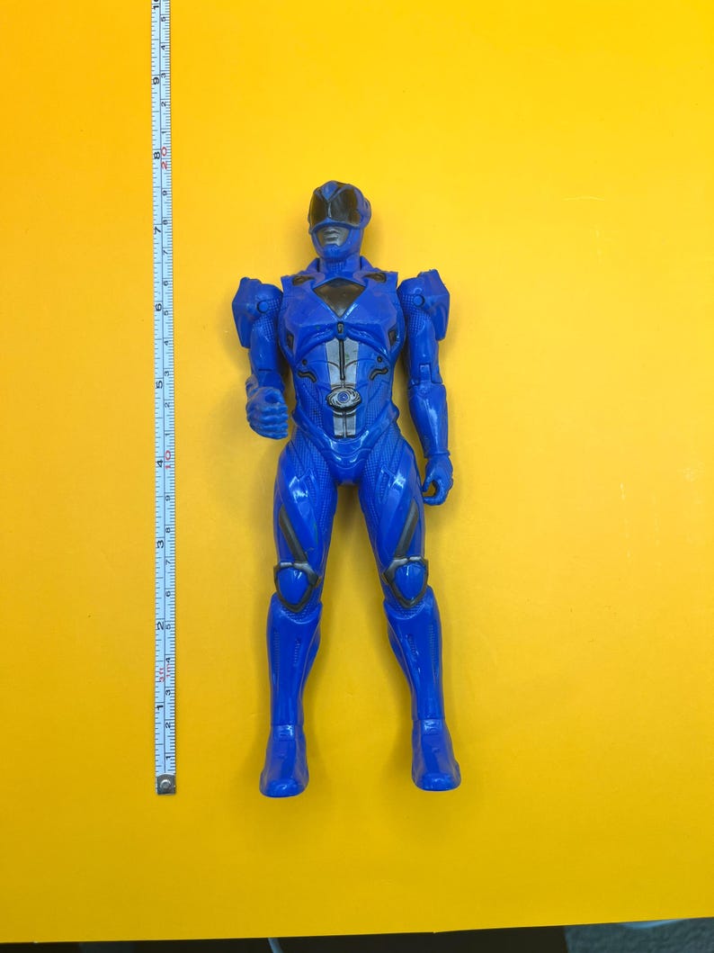 May include: A blue superhero action figure, standing on a yellow surface. The figure is approximately 25 cm tall, with detailed armor and a helmet. A measuring tape is positioned next to the figure.