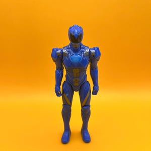 May include: A blue action figure of a superhero with a helmet and armour. The figure has silver and gold accents and is posed standing against a yellow background. The figure is a Power Ranger.