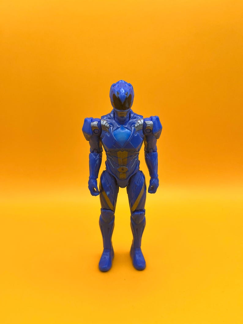 May include: A blue action figure of a superhero with a helmet and armour. The figure has silver and gold accents and is posed standing against a yellow background. The figure is a Power Ranger.