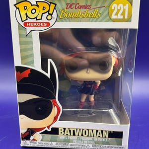 May include: A Batwoman vinyl figure from the DC Comics Bombshells series, number 221, is displayed in its original packaging. The figure is in a dynamic pose, wearing a black and red costume. The packaging features the Pop! Heroes logo.