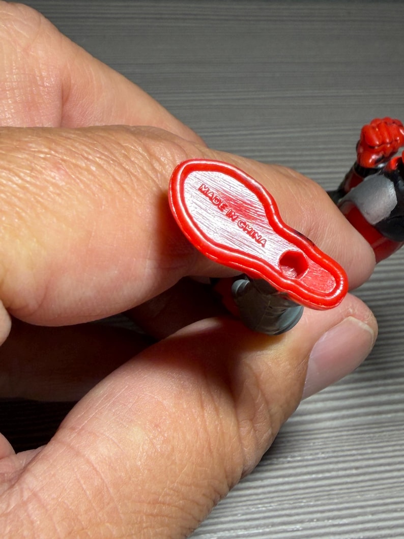 May include: A red plastic foot of an action figure, with the words "MADE IN CHINA" printed on the sole. The figure's leg is gray and black. The figure is held by a person.