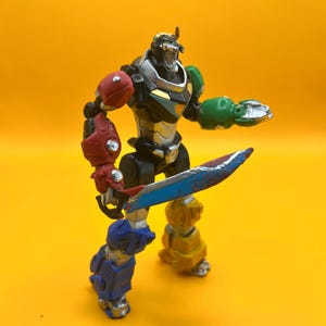 May include: A colorful action figure of a robot warrior, posed against a yellow background. The robot has a silver head, black torso, and limbs in red, green, blue, and yellow. It holds a blue and red sword.