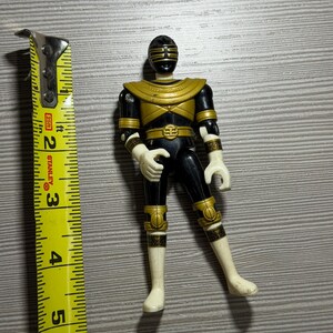 May include: A black and gold Power Ranger action figure, posed with one leg bent. The figure is approximately 13 cm tall, with gold detailing on the chest, belt, and limbs. The figure has a black helmet with gold detailing.
