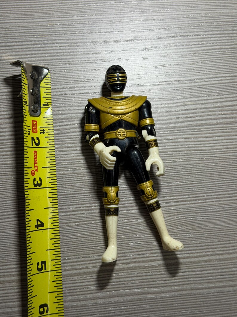 May include: A black and gold Power Ranger action figure, posed with one leg bent. The figure is approximately 13 cm tall, with gold detailing on the chest, belt, and limbs. The figure has a black helmet with gold detailing.