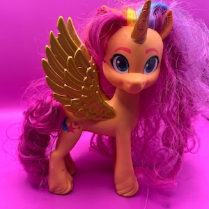 May include: A toy pony with a peach-colored body, gold wings, and a golden horn. The pony has long, curly pink and purple hair. It has blue eyes and a star and crescent moon tattoo on its leg. The pony is standing on a pink surface.