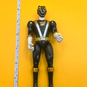 以下が含まれることがあります： 金と白のアクセントが付いた黒いアクションフィギュア。フィギュアは白い頭と手、金のベルト、ブーツと腕のアクセントがあります。黄色い背景に立っています。