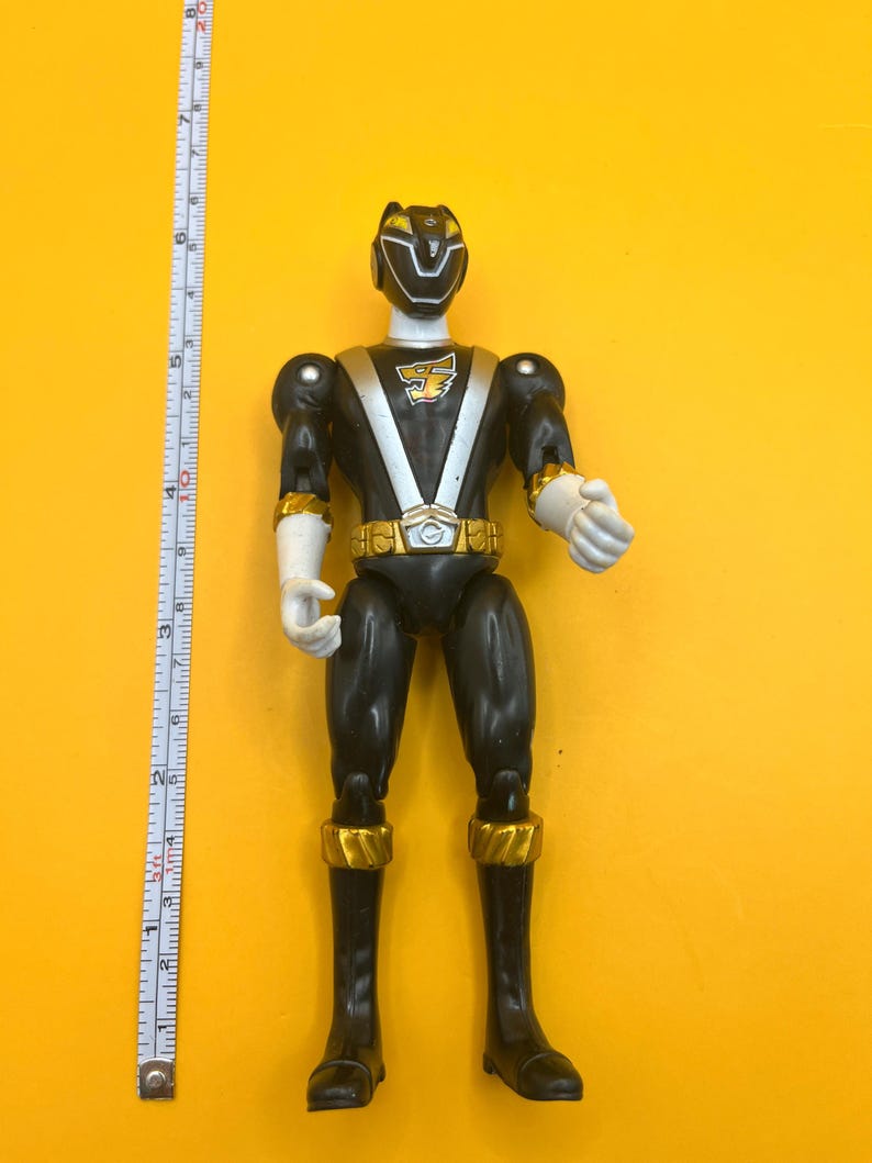 以下が含まれることがあります： 金と白のアクセントが付いた黒いアクションフィギュア。フィギュアは白い頭と手、金のベルト、ブーツと腕のアクセントがあります。黄色い背景に立っています。