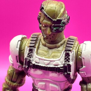 K&ouml;nnte beinhalten: Nahaufnahme einer milit&auml;rischen Actionfigur. Die Figur ist haupts&auml;chlich olivgr&uuml;n mit wei&szlig;en und schwarzen Akzenten. Sie hat einen Helm mit Visier, eine Gesichtsmaske und eine taktische Weste. Der Hintergrund ist ein kr&auml;ftiges Pink.