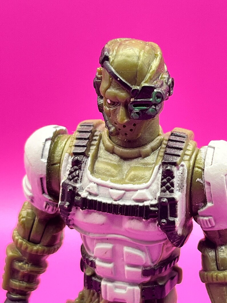 K&ouml;nnte beinhalten: Nahaufnahme einer milit&auml;rischen Actionfigur. Die Figur ist haupts&auml;chlich olivgr&uuml;n mit wei&szlig;en und schwarzen Akzenten. Sie hat einen Helm mit Visier, eine Gesichtsmaske und eine taktische Weste. Der Hintergrund ist ein kr&auml;ftiges Pink.