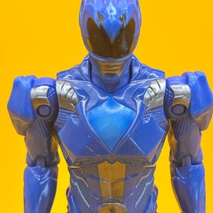 May include: A blue action figure of a superhero with a helmet and armour. The figure has silver accents on the shoulders and chest. The background is a solid yellow.