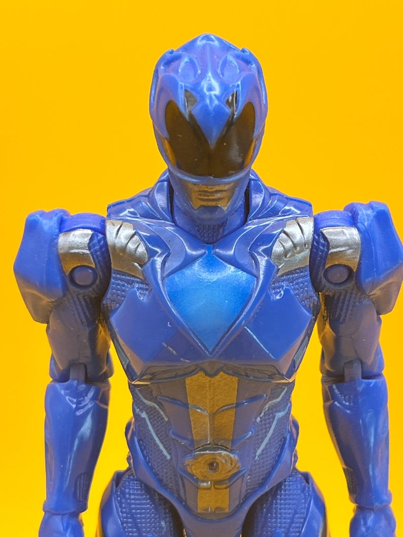 May include: A blue action figure of a superhero with a helmet and armour. The figure has silver accents on the shoulders and chest. The background is a solid yellow.