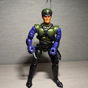 May include: A detailed action figure of a humanoid insect-like character. The figure features a green helmet, chest armor, and black and purple accents. The figure has red eyes and is posed with arms at its sides.