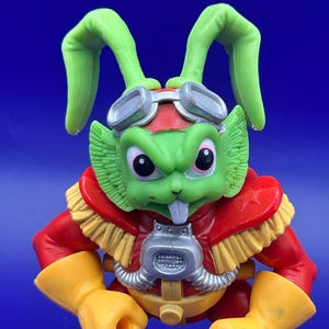 May include: A green rabbit-like action figure with large ears, wearing goggles, a red and yellow uniform, and yellow gloves. The figure has a determined expression and is posed against a blue background. The toy is a collectible item.