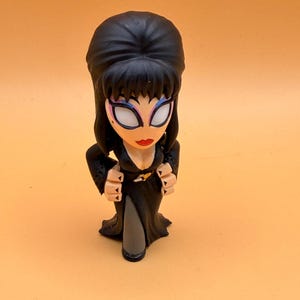 May include: A figurine of a woman with black hair, a black dress, and a plunging neckline. The figurine has a pale face with red lipstick and dramatic eye makeup. The figure is posed with hands clasped near the chest.