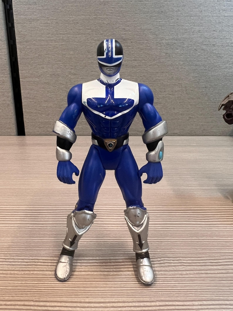 May include: A blue action figure of a Power Ranger, standing upright. The figure has a blue and white uniform with silver accents on the arms and legs. The figure has a black helmet with white details and a gold belt buckle.