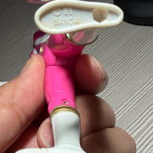 May include: A pink and white action figure of a Power Ranger. The figure has gold accents and white boots. The bottom of the foot reads "MADE IN CHINA". The figure is held by a person.