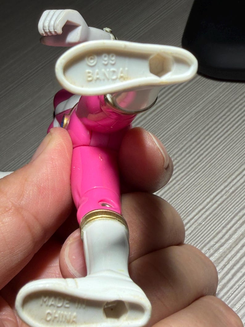 May include: A pink and white action figure of a Power Ranger. The figure has gold accents and white boots. The bottom of the foot reads "MADE IN CHINA". The figure is held by a person.