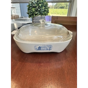 Pode incluir: Travessa branca com tampa de vidro transparente. A travessa tem um desenho de casa de fazenda azul e branco. A tampa tem um botão de vidro transparente. A travessa está sobre uma mesa de madeira.