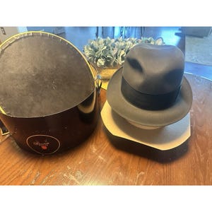 May include: A dark gray fedora hat with a black band, resting on a hat stand. The hat is next to a black hat box with a gold rim and the text "Churchill". The hat box is open, revealing a black interior.