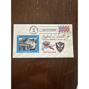 Puede incluir: Un sobre con un sello de "Primer día de emisión", matasellado en Kansas City, MO, el 16 de agosto de 1999. Presenta un sello azul con una foto y el texto "Honoring Those Who Served". El sobre también incluye la firma del coronel Robert L. Scott Jr. y emblemas del 23rd Fighter Group.