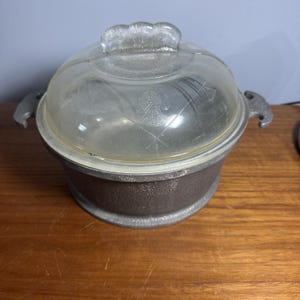 May include: A round, vintage casserole dish with a clear glass lid and a metal base. The lid has a decorative handle and etched design. The dish has two side handles. The base is a dark, textured metal.