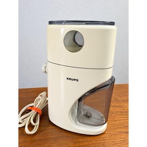 May include: A cream-colored electric coffee grinder with a rounded rectangular body, a dark gray transparent bean hopper lid, a matching dark gray grounds container, and a side-mounted dial. The brand name KRUPS is printed in black on the front.