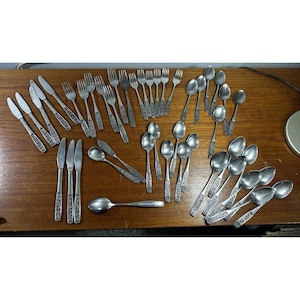 May include: A large collection of silver-toned stainless steel flatware, including various knives, forks, and spoons. Each piece features a textured, decorative pattern on the handle. The set is arranged in groups on a flat surface.