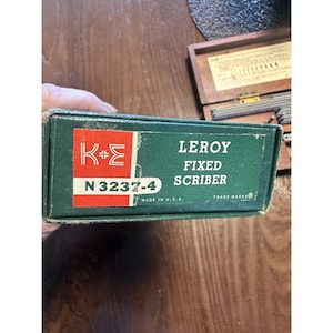 Peut inclure: Une boîte verte avec le texte "LEROY FIXED SCRIBER" et le logo K+E. La boîte porte également la mention "N 3237-4" et "MADE IN U.S.A.". La boîte est ouverte, révélant un ensemble d'outils.