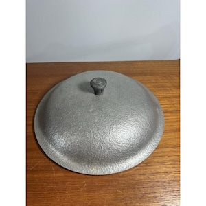 May include: A round, gray metal lid with a textured surface and a small knob on top. The lid is likely for a pot or pan and is made of a durable material. The lid is sitting on a wooden surface.