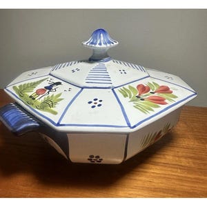 Könnte beinhalten: Eine weiße Keramik-Servierschüssel mit Deckel, verziert mit handgemalten Blumen- und geometrischen Designs in Rot, Blau und Grün. Die Schüssel hat eine achteckige Form und einen blauen Griff.