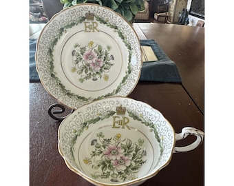 Könnte beinhalten: Ein Vintage-Tee-Set, bestehend aus einer Tasse und einer Untertasse. Das weiße Porzellan hat ein Blumendesign mit rosa Blüten und grünen Blättern. Der Rand ist gewellt und mit Gold verziert. Die Buchstaben "ER" und eine Krone sind vorhanden.