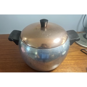 Könnte beinhalten: Ein Vintage-Eiskübel mit kupferfarbenem Deckel und silberfarbenem Körper. Der Eiskübel hat zwei schwarze Griffe und einen schwarzen Knopf auf dem Deckel. Der Eiskübel steht auf einer Holzoberfläche.