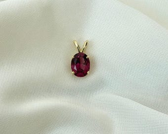 May include: A gold pendant featuring an oval-cut deep red gemstone secured by four prongs. The pendant has a simple V-shaped bail at the top, designed to hold a chain. The gemstone displays a faceted surface that reflects light with a dark crimson hue.