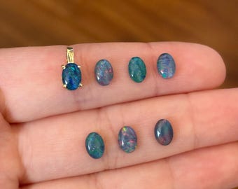 May include: Seven oval-shaped black opals, some with iridescent colors, are displayed on a hand. One opal is on a gold chain.