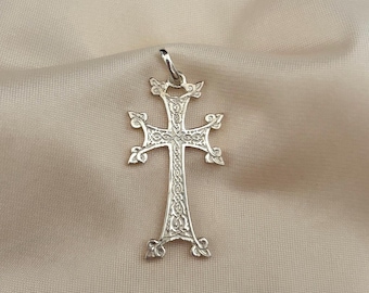 May include: A silver pendant in the shape of a cross, featuring intricate filigree detailing. The cross has ornate embellishments at the ends of each arm and at the top and bottom. The pendant hangs from a small loop and is displayed against a neutral fabric.