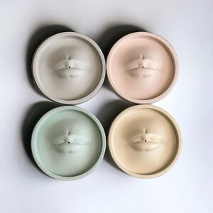 May include: Four round concrete incense holders with a raised lip and a small hole in the centre. Each holder features a different colour: light grey, light pink, light green, and light beige. The holders have a 3D design of a pair of lips.
