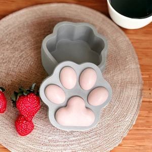 Könnte beinhalten: Eine graue Beton-Schmuckschatulle in Form einer Pfote mit einem abnehmbaren Deckel. Der Deckel ist geöffnet und das Innere der Schachtel ist sichtbar. Die Schachtel hat ein pinkfarbenes Pfotenmuster auf dem Boden.