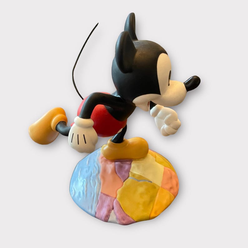 May include: A Mickey Mouse figurine in a dynamic pose, with black ears, a white face, and a red shorts. The figure is balanced on a colorful, patchwork-style base. The figurine has yellow shoes and white gloves.