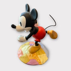 May include: A Mickey Mouse figurine in a dynamic pose, featuring a black head and ears, white face, red shorts, and yellow shoes. The figure is balanced on a colorful, patchwork-style base. The figurine's tail is a thin, black wire.