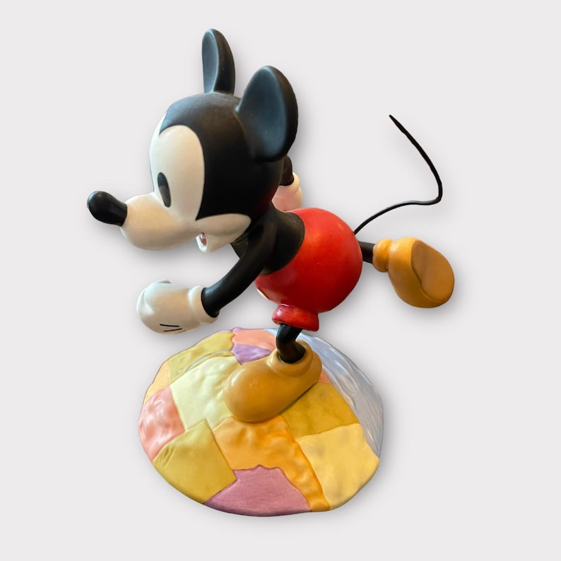 May include: A Mickey Mouse figurine in a dynamic pose, featuring a black head and ears, white face, red shorts, and yellow shoes. The figure is balanced on a colorful, patchwork-style base. The figurine's tail is a thin, black wire.