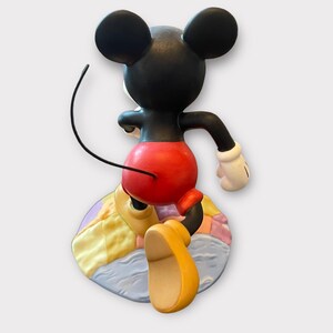 May include: A figurine of Mickey Mouse from the back, with black ears and head, red shorts, yellow shoes, and white gloves. The figure is posed on a colorful, circular base with pastel colors.