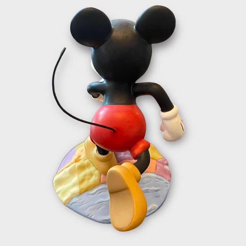 May include: A figurine of Mickey Mouse from the back, with black ears and head, red shorts, yellow shoes, and white gloves. The figure is posed on a colorful, circular base with pastel colors.