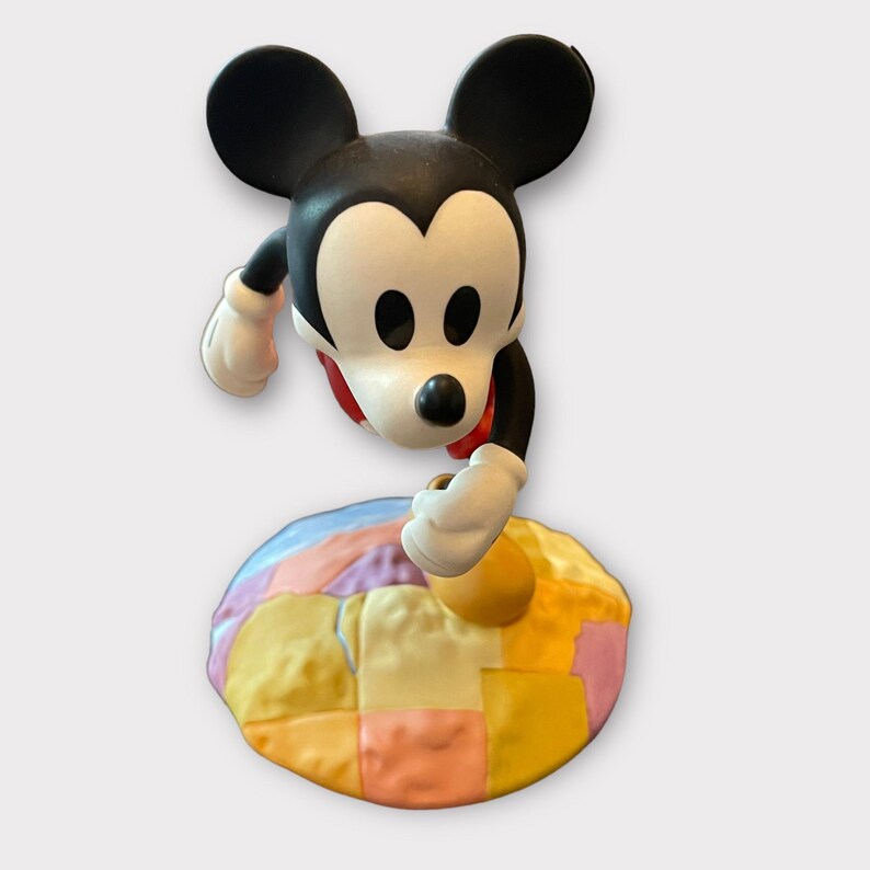 May include: A Mickey Mouse figurine with black ears, a white face, and a red bow tie. The figure is positioned above a colorful, patchwork-style base with pastel hues of yellow, pink, and blue. The figurine is a collectible item.