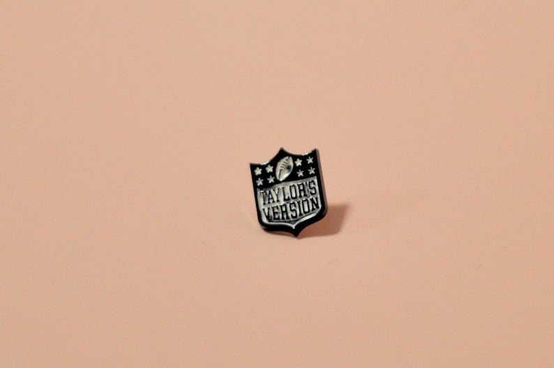 K&ouml;nnte beinhalten: Schwarzer und wei&szlig;er Emaille-Pin in Form eines Fu&szlig;ballschilds mit dem Text "Taylor's Version" im Inneren.