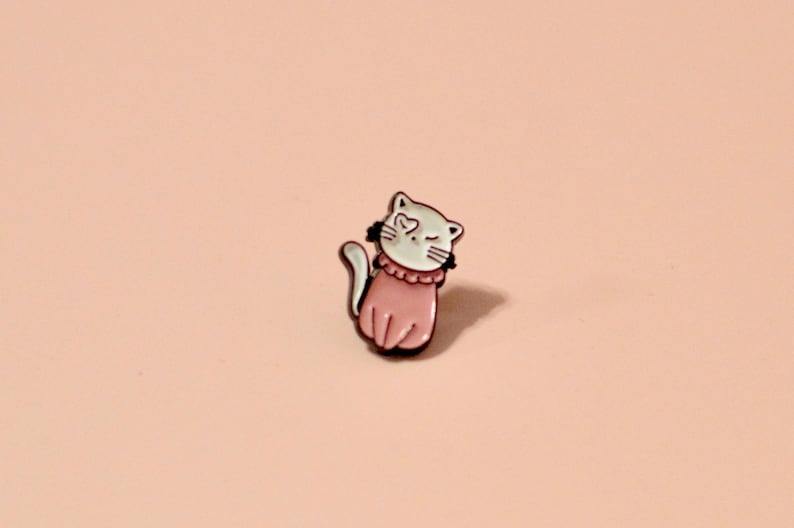 K&ouml;nnte beinhalten: Eine Emaille-Anstecknadel mit einem Cartoon-Katzenmotiv, das ein rosa Kleid tr&auml;gt. Die Katze zwinkert und hat ein herzf&ouml;rmiges Auge.
