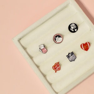 K&ouml;nnte beinhalten: Ein wei&szlig;es Samttablett mit sechs Emaille-Anstecknadeln. Die Nadeln zeigen verschiedene Designs, darunter eine Katze, ein Herz, ein Fu&szlig;ball und ein Frauen Gesicht.